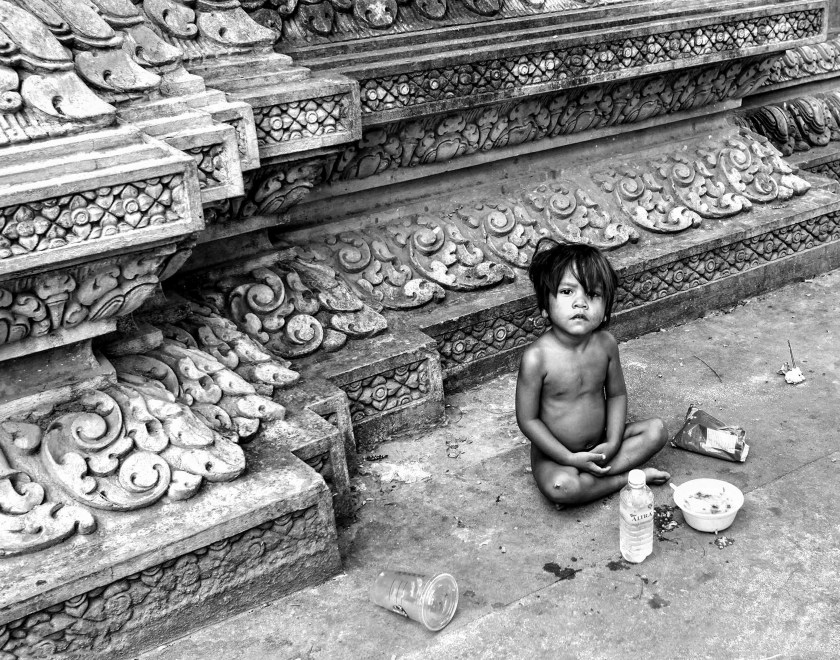 A young child sitting on the ground beside ornate stone carvings, with a bottle of water and some food scraps nearby, in a black and white setting.
