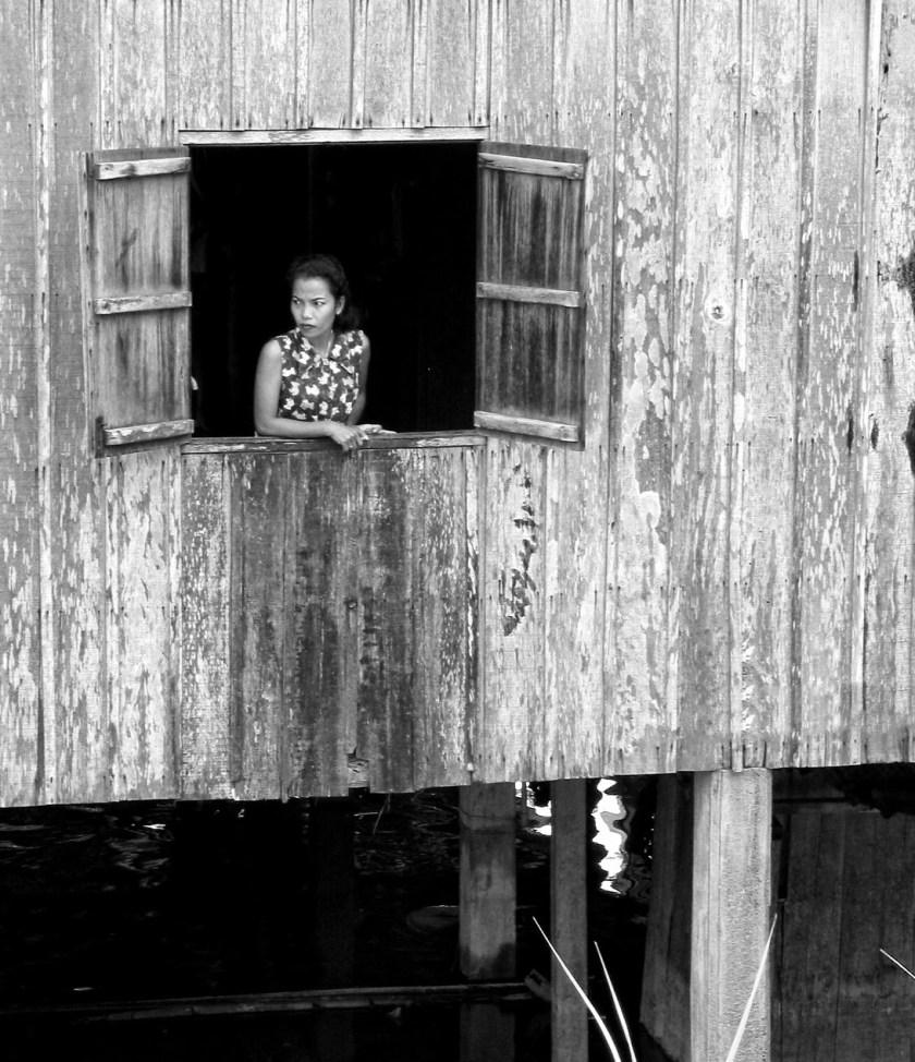 A young woman gazes thoughtfully out of a window of a wooden house, surrounded by peeling paint and weathered wood. The image is in black and white.