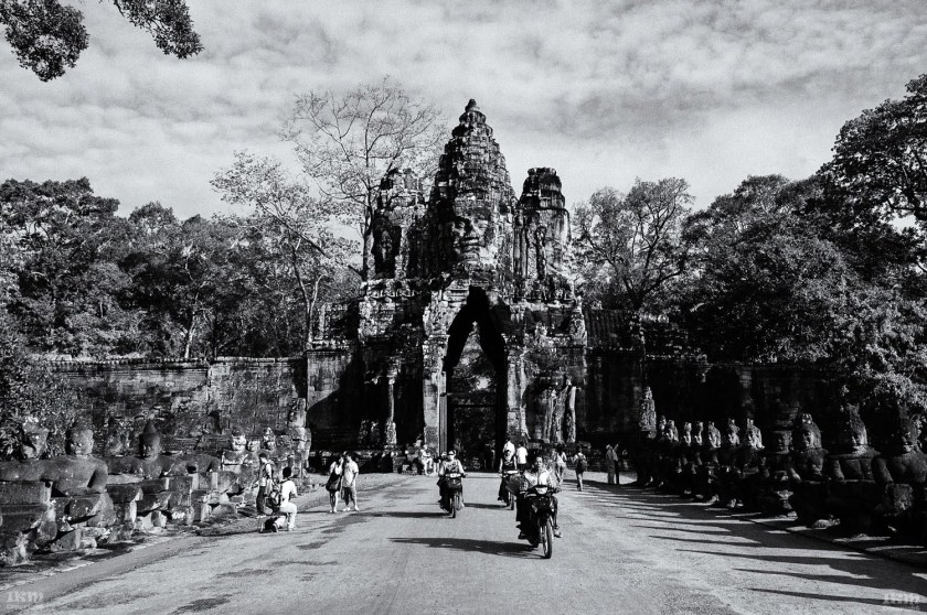 Black and white photograph of an ancient stone gate with intricately carved faces, surrounded by trees, with people walking and motorcycles passing underneath.