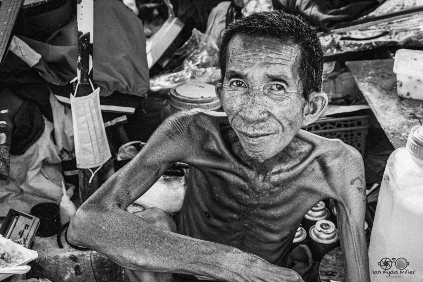 A thin, elderly man sitting amidst clutter, with visible signs of wear on his skin, smiling gently at the camera.