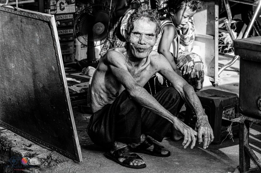 An elderly man sitting on the ground with a slight smile, wearing black pants and sandals, in a cluttered environment. A child can be seen in the background.