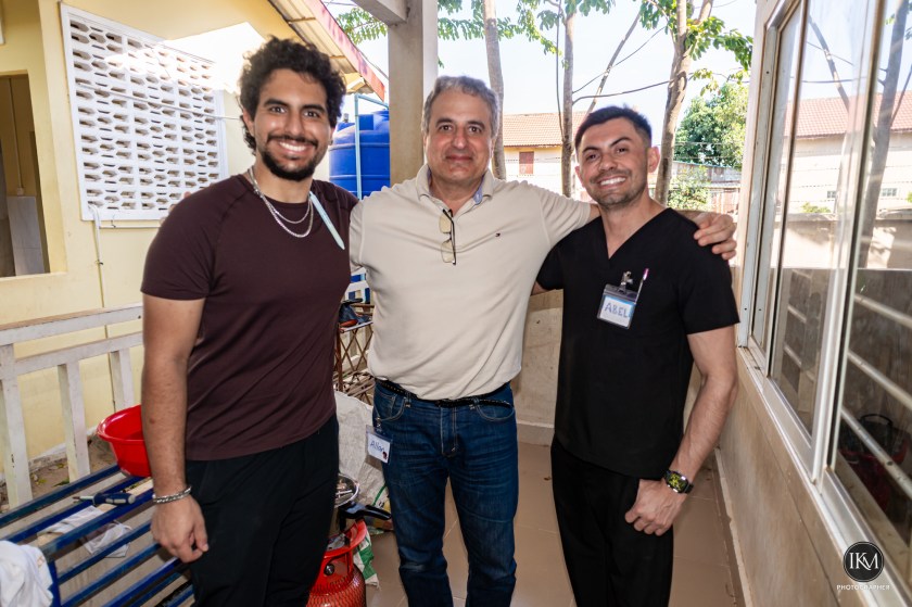 Three men posing together in a casual outdoor setting, with trees and a building in the background. One man is wearing a dark shirt and jeans, another in a light-colored sweater and glasses, and the third in a black medical scrub with a name tag.