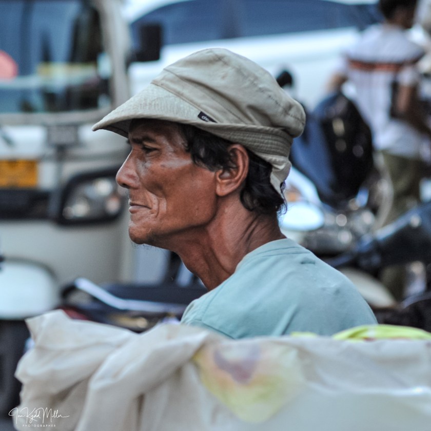 A side profile of an elderly man wearing a light-colored bucket hat and a blue shirt, with a subtle smile and wrinkles on his face, set against a backdrop of blurred vehicles.