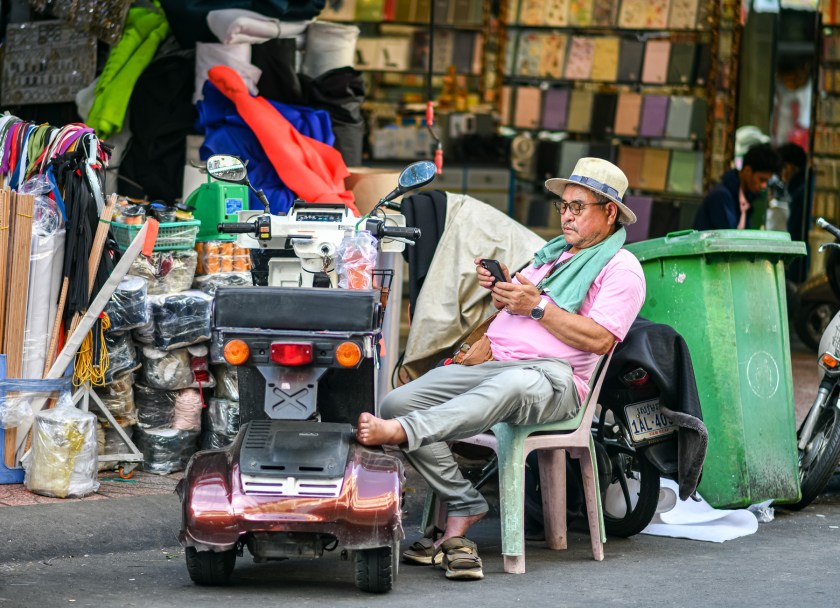 An older man sitting on a plastic chair, using a smartphone, next to a small electric scooter. He is wearing glasses, a pink shirt, and a straw hat. Behind him is a colorful array of fabric and materials, along with a green trash bin.