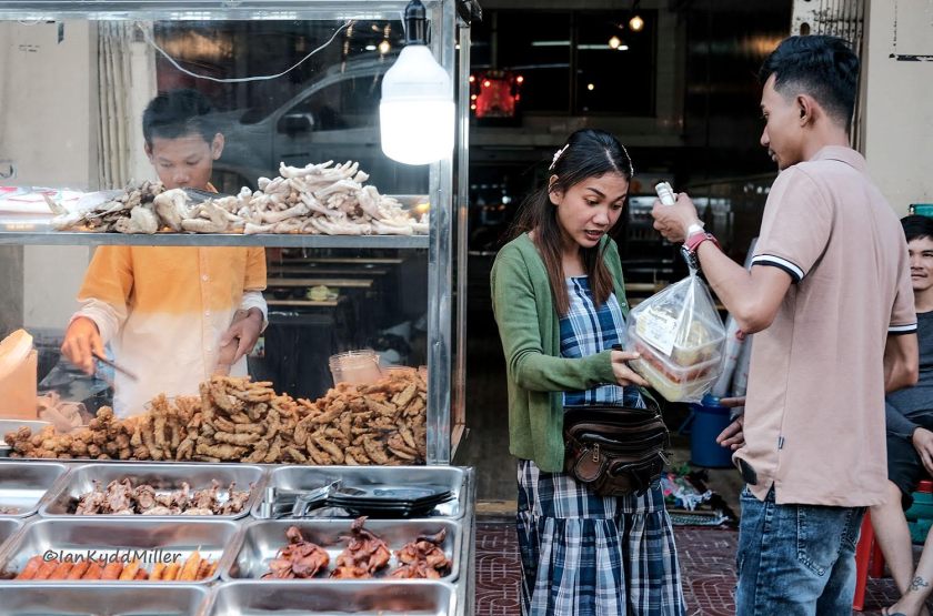 A street food vendor offers various fried snacks in a glass display, while a woman receives a takeaway order from a man. They are engaging in conversation, with other customers in the background.