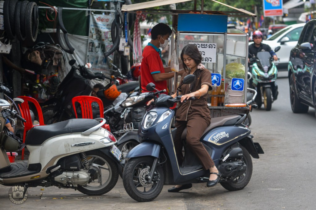 A young woman in brown clothing is seated on a blue scooter while using a phone, parked near a food stall with a vendor in a red shirt. Various motorcycles and a busy street can be seen in the background.
