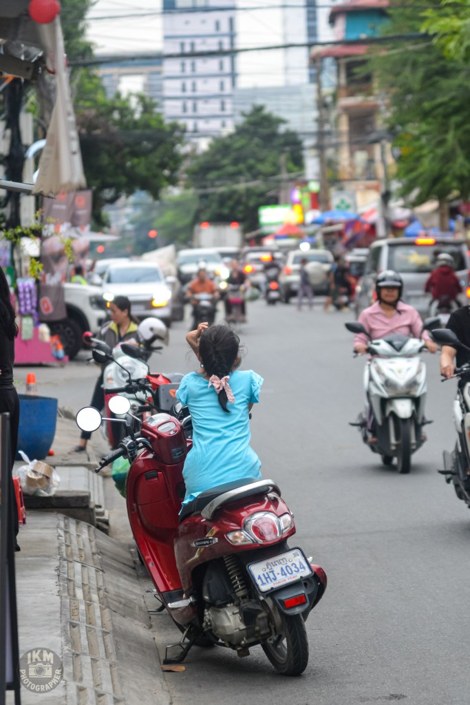 A girl wearing a blue dress sitting on a red motorcycle parked on the street, with traffic and buildings in the background.