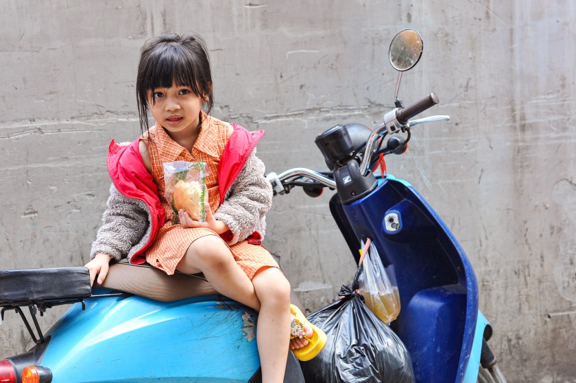 A young girl sitting on a blue motorcycle, holding a snack in a plastic bag, wearing a red jacket over a checkered dress.