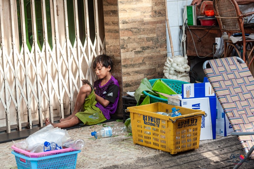 A young boy sitting on the ground against a wall, looking at a smartphone, surrounded by plastic waste and boxes.