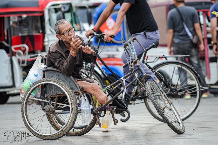 An elderly man in a wheelchair bicycle sits on a city street, gesturing with his hands while surrounded by pedestrians and bicycles.