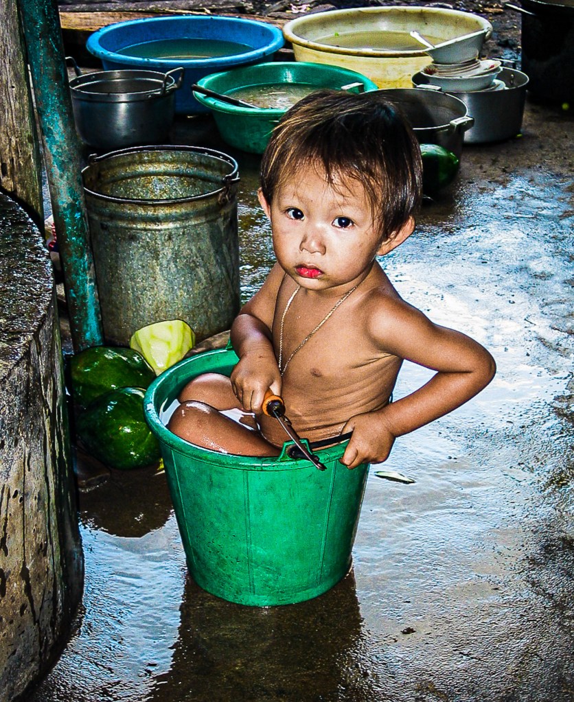 A young child sitting in a green bucket, surrounded by various containers and water on the floor.