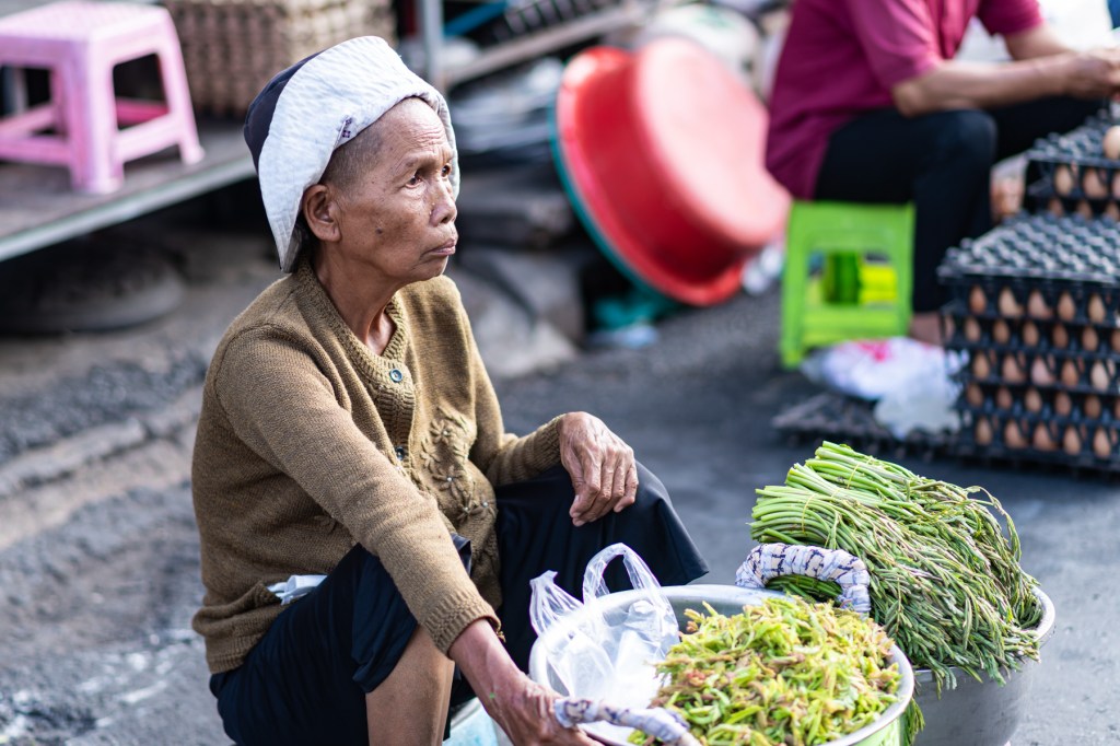 An elderly woman sitting at a market with vegetables, wearing a white headscarf and a brown cardigan.