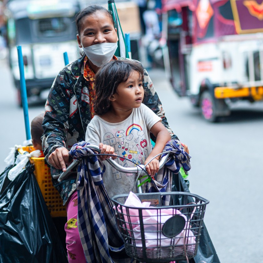 A smiling woman wearing a mask rides a bicycle with a young girl in a city street, showcasing a colorful backdrop and urban life.