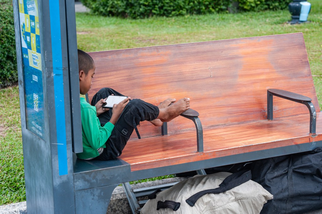 A young boy sitting on a bus stop bench, focused on using a mobile device, with his bare feet resting on the ground.