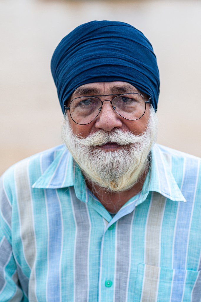 Portrait of an older man with a white beard and glasses, wearing a blue turban and a striped shirt.