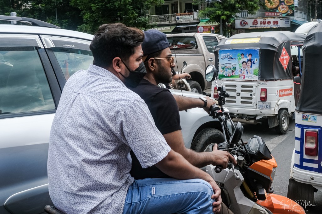 Two men riding a motorcycle in traffic, one seated in front and the other behind, both wearing masks.