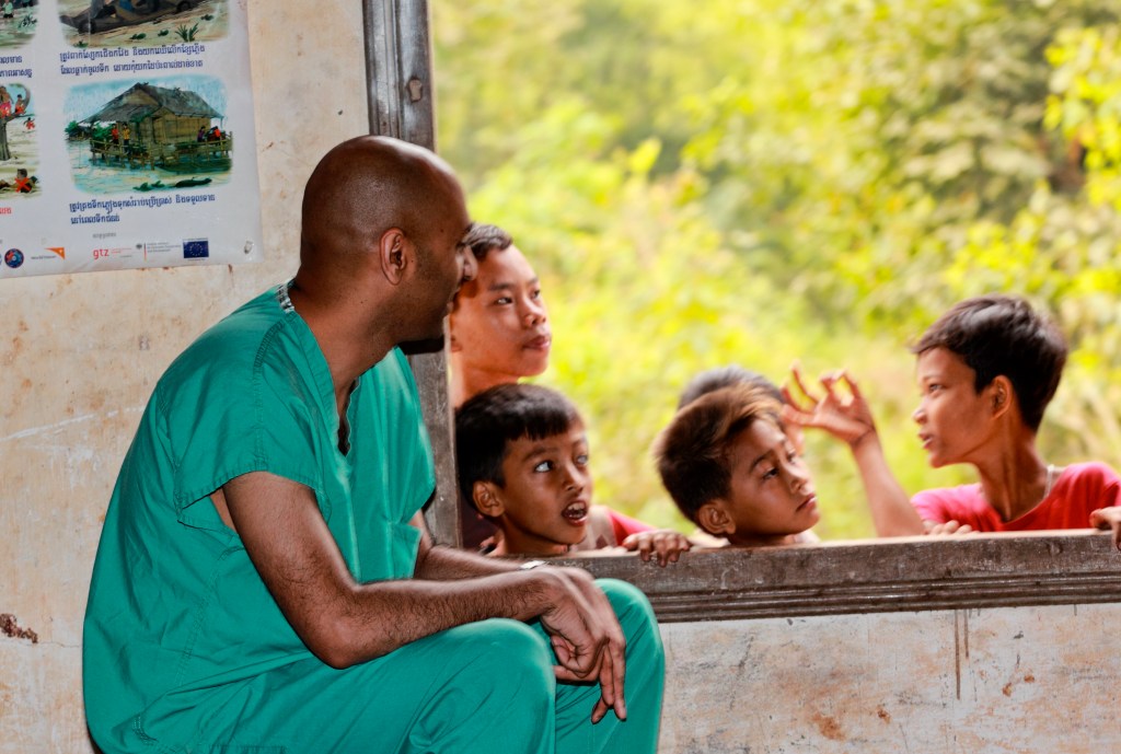A healthcare worker in green scrubs sits indoors, looking out a window at four children gathered outside, engaged in conversation.