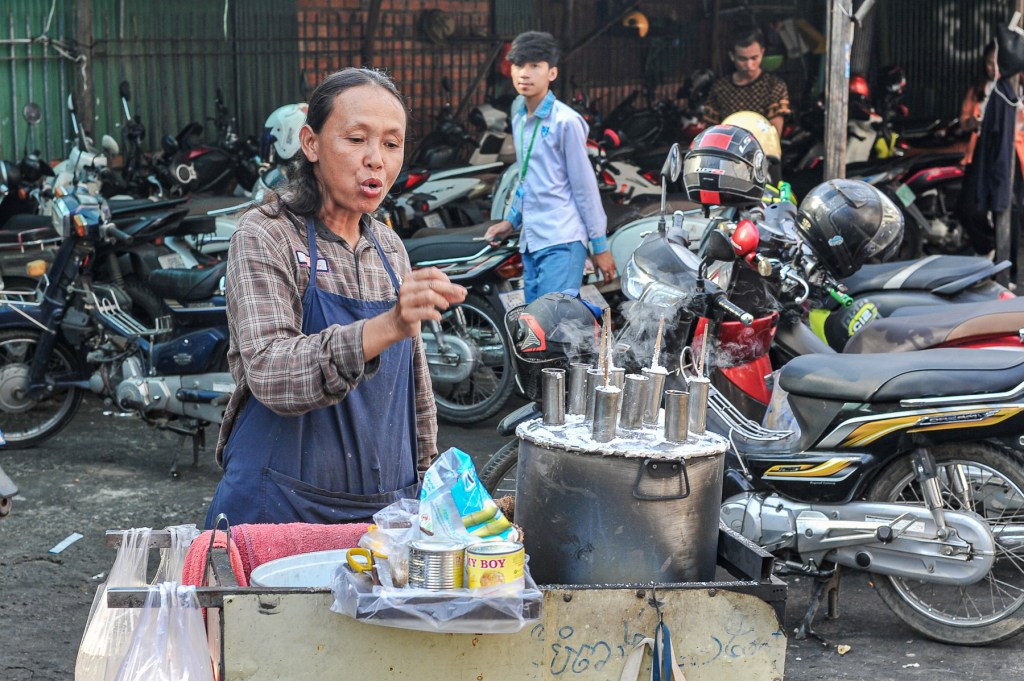 A street vendor in a blue apron prepares food with steaming pots in front of a variety of parked motorcycles.