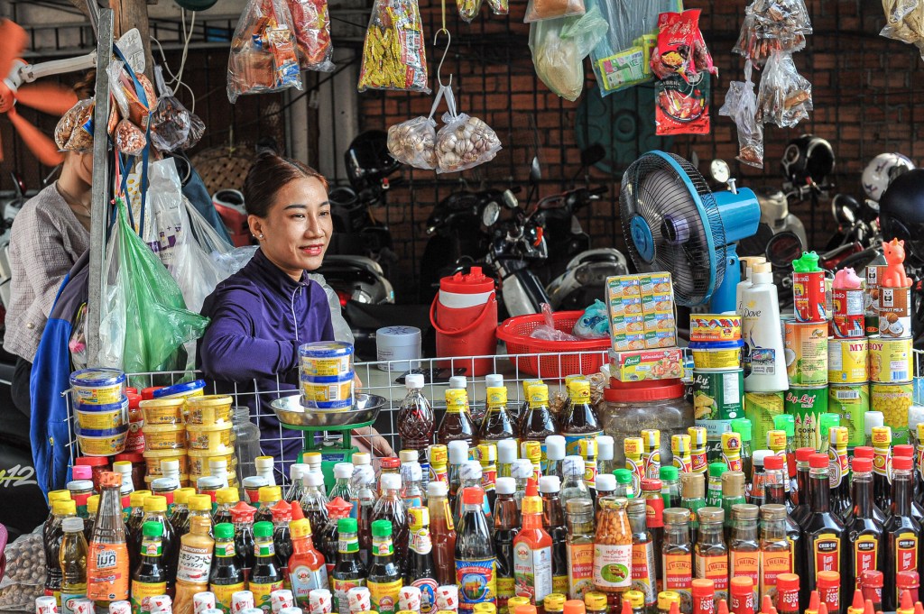 A market vendor smiles while seated at her stall, surrounded by various condiments, sauces, and snacks. In the background, other stall items hang and there are motorcycles parked.