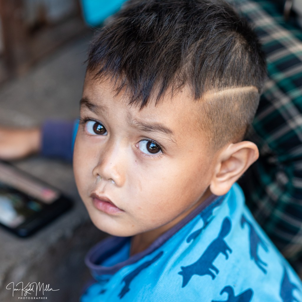 Close-up portrait of a young boy with short hair and a patterned blue shirt, looking directly at the camera with a serious expression.