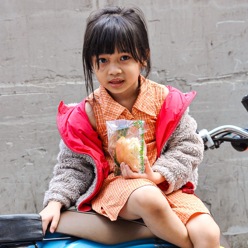 A young girl sitting on a bicycle wearing a red jacket over an orange checkered dress, holding a packaged snack.