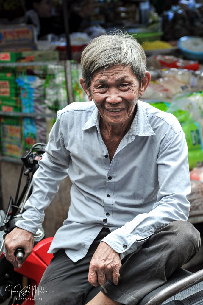 An elderly man with gray hair smiling while sitting on a motorcycle in a market setting.