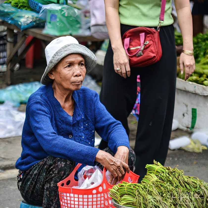 An elderly woman sitting on the ground in a market, wearing a blue sweater and a white hat, holding a red basket of goods, with fresh vegetables in front of her and shoppers in the background.