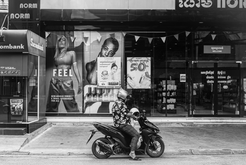 A person wearing a helmet sits on a motorbike in front of a storefront displaying advertisements for fragrances and promotional discounts. The image is in black and white.