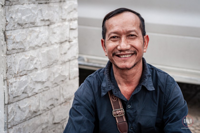 A smiling man sitting in front of a textured stone wall, wearing a dark shirt and a shoulder bag.