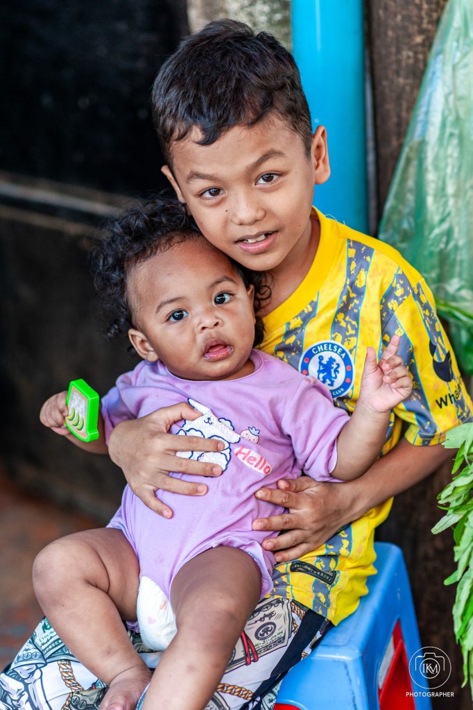 A young boy holding a baby, both smiling at the camera. The boy is wearing a yellow and blue striped shirt, while the baby is dressed in a light purple onesie. They are seated on a small blue stool with greenery visible in the background.