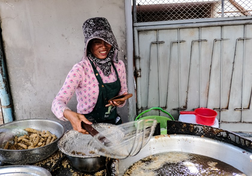 A woman wearing a hat and apron smiles while cooking at a street food stall, frying food in a large pan.