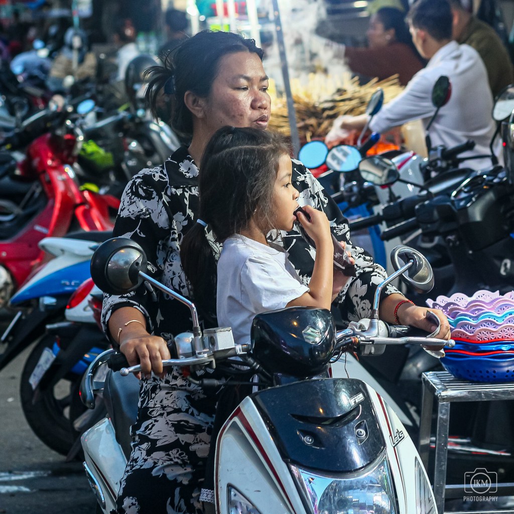 A woman riding a motorcycle with a child sitting in front of her, both in a bustling market setting surrounded by parked scooters.