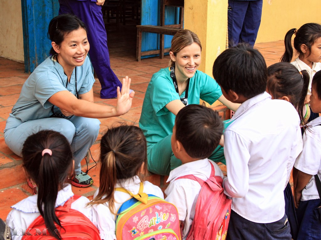 Two healthcare professionals interact with children at a school, smiling and engaging in a cheerful conversation.