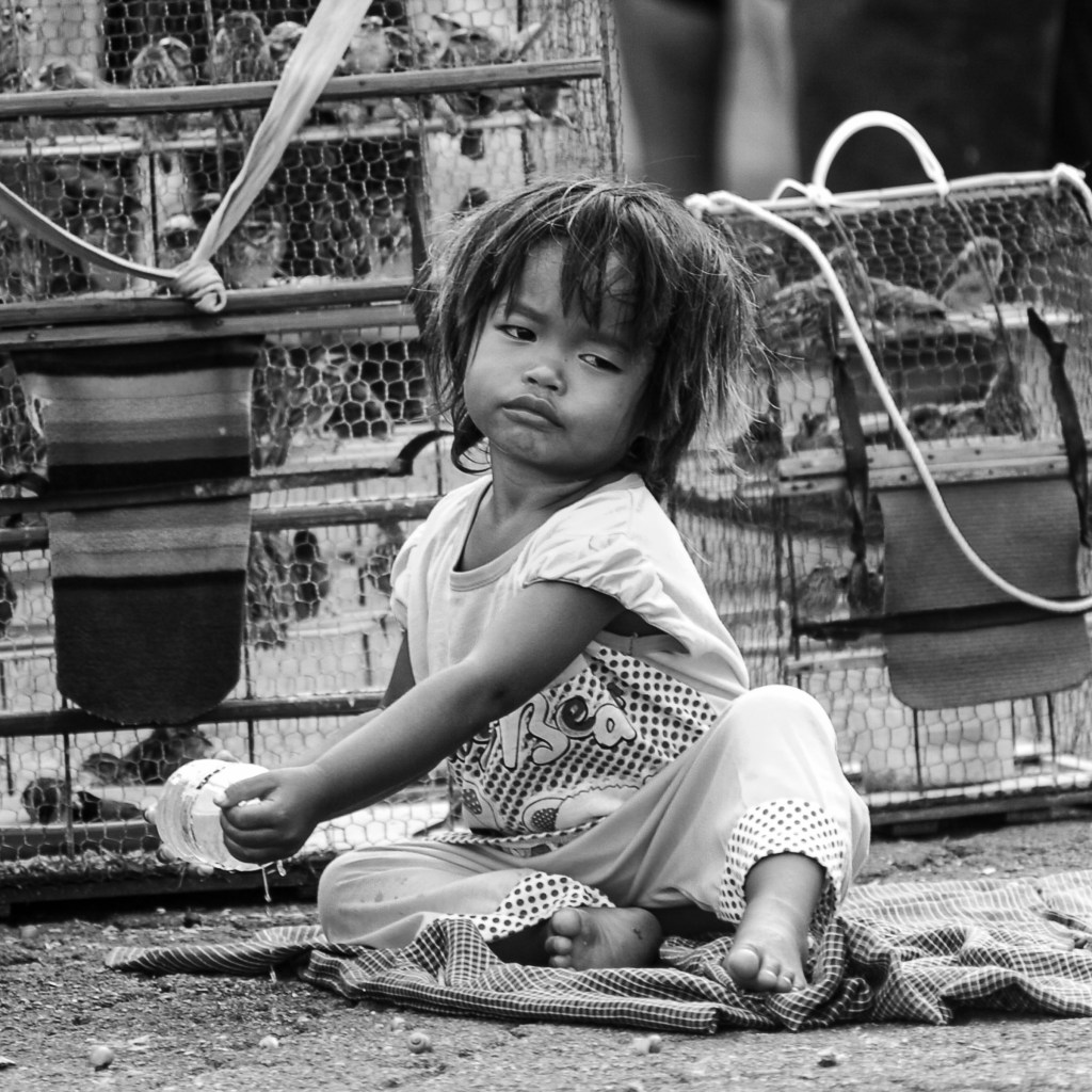 A young child sitting on the ground with a serious expression, holding a container, surrounded by cages in a black and white setting.