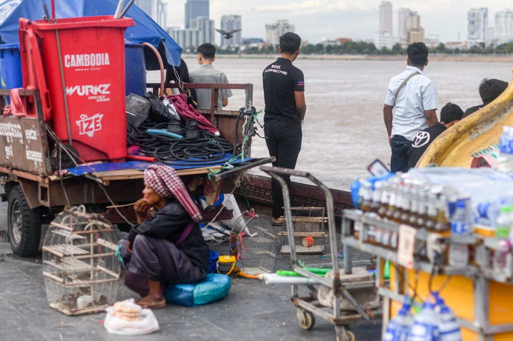 A scene at a riverside with a red cart labeled 'Cambodia' and various items, including drinks and equipment. A woman sits on the ground with a bird cage, while several people stand nearby overlooking the river and city skyline.