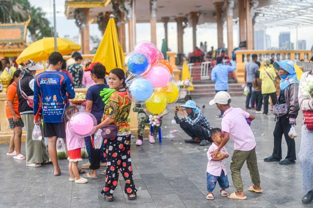 A bustling outdoor scene featuring a woman holding colorful balloons, surrounded by people engaging in various activities in a lively atmosphere.