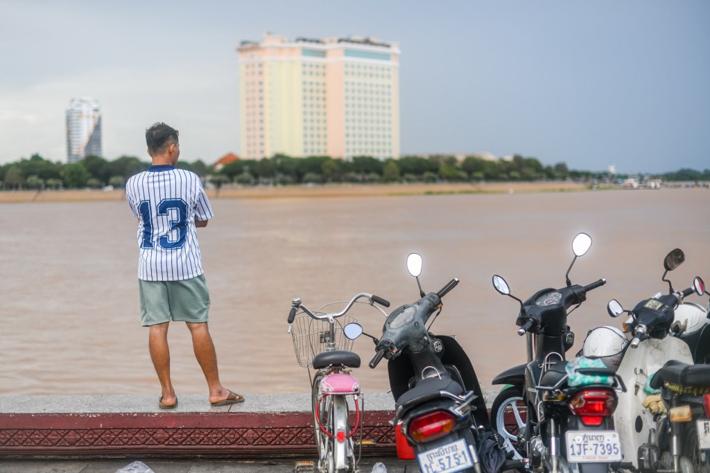 A person in a striped jersey and shorts stands by a river, overlooking the water. Several parked motorcycles are lined up nearby, and buildings are visible in the background under a cloudy sky.