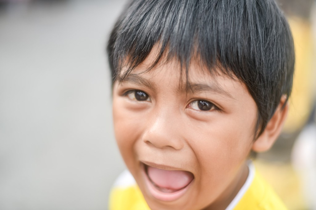 A close-up of a young boy with short black hair, smiling widely and showing excitement.