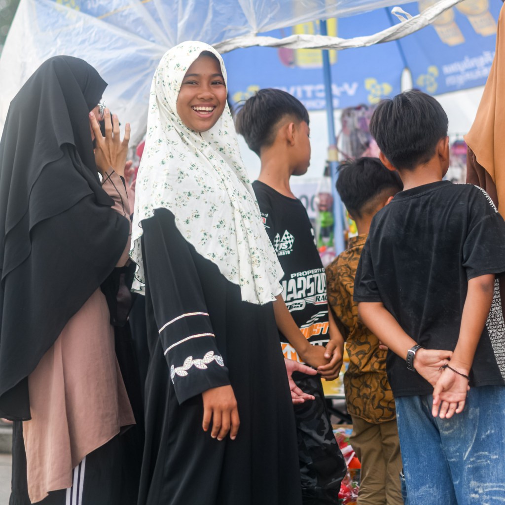 A group of children and young adults standing together at a market, with one girl in the foreground wearing a white hijab with floral patterns and smiling happily. Others are partially visible, engaged in conversation and looking at various items.