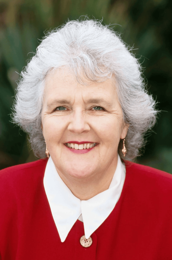 A smiling older woman with gray hair, wearing a red jacket and white collar, poses for a portrait against a blurred natural background.