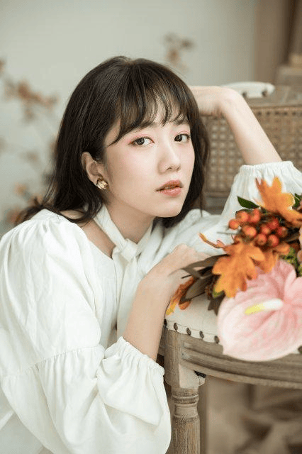 A young woman with long dark hair, wearing a white blouse, rests her chin on her hand while posing with a bouquet of flowers, including orange and pink blooms, in a soft, aesthetic setting.