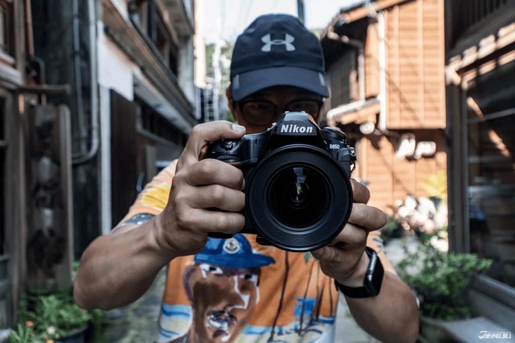 A person holding a Nikon D850 camera and pointing it towards the viewer, dressed in a colorful shirt, with a cityscape in the background.