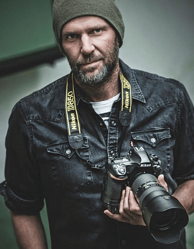A male photographer wearing a beanie and denim shirt, holding a Nikon camera with a large lens.