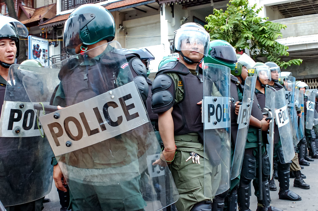 A line of police officers in riot gear, holding shields labeled 'POLICE'. They are standing in formation, ready for crowd control.