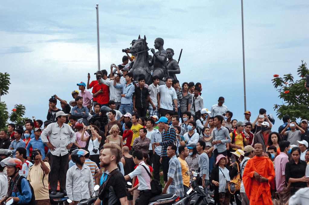 A large crowd of people gathered around a statue, with some individuals taking photos. The crowd is diverse, wearing various clothing styles and hats, under a cloudy sky.