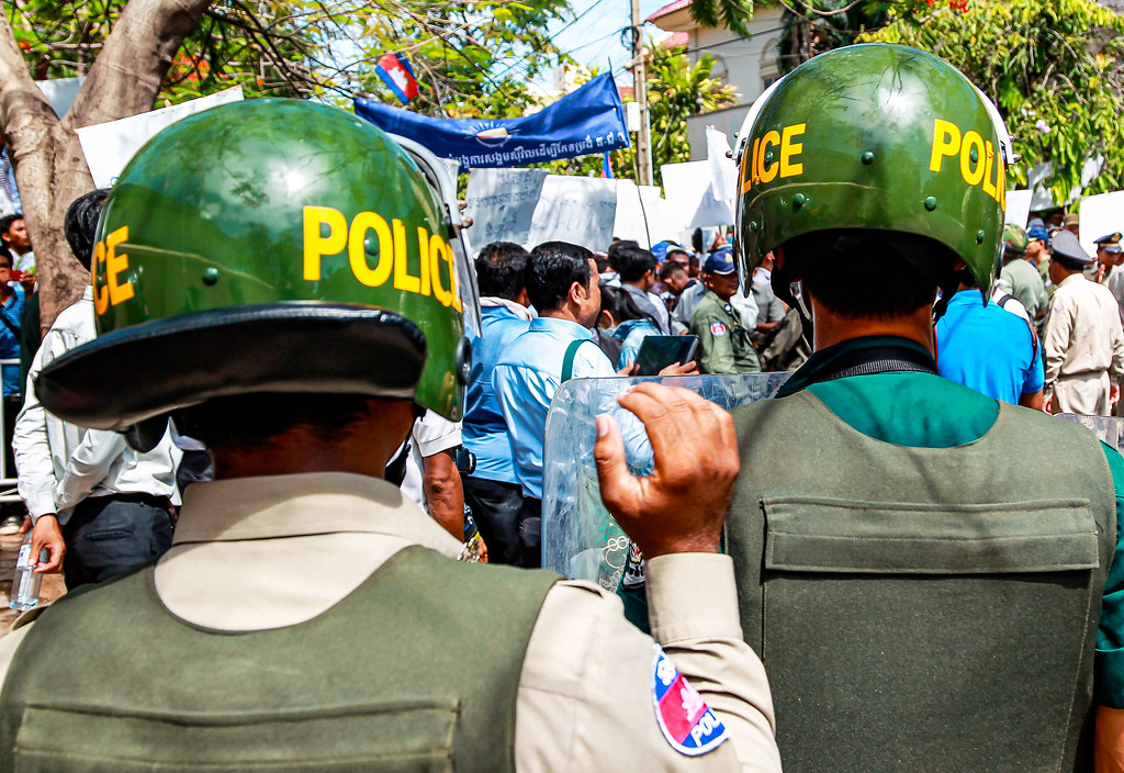 Two police officers in helmets stand facing a crowd during a protest, with banners and signs visible in the background.