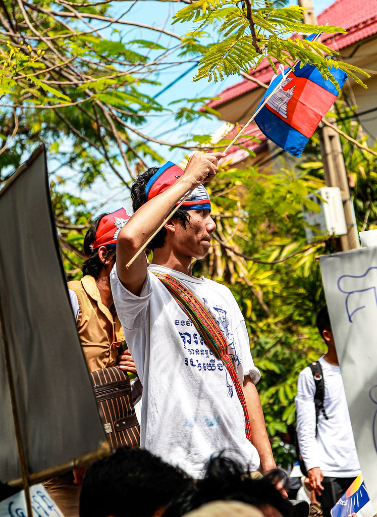 A young man holding a Cambodian flag, wearing a white t-shirt and a colorful sash, surrounded by greenery at a demonstration.