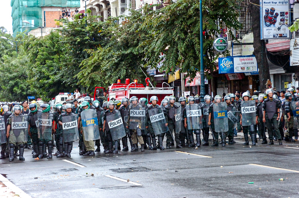 A line of police officers in riot gear standing on a street, with shields marked 'POLICE', amidst a city backdrop with trees and buildings.