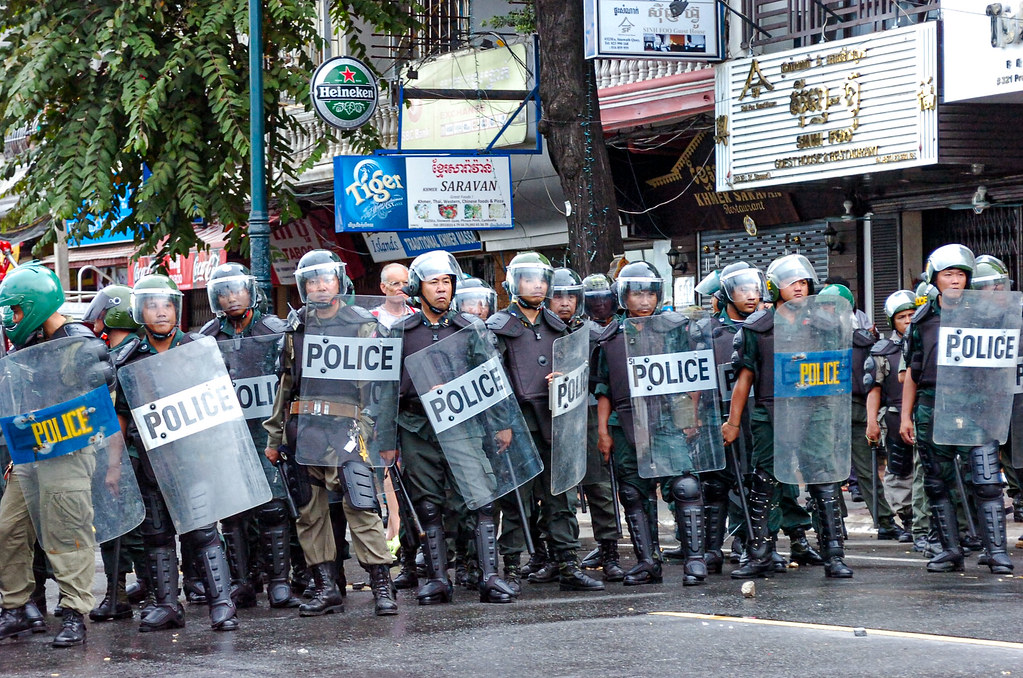 A line of police officers in riot gear holding shields labeled 'POLICE' during a protest or disturbance in an urban setting.