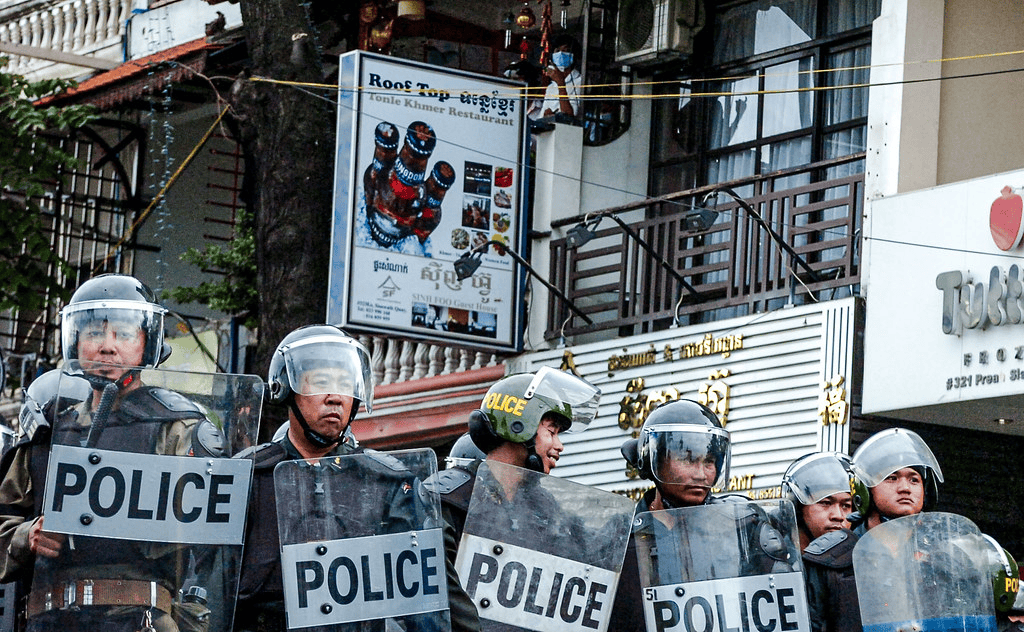 A line of police officers in riot gear holding shields with the word 'POLICE' displayed, standing in front of a storefront with a sign for a restaurant in the background.